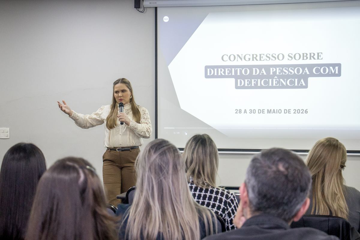 A Dra. Priscilla Machado palestrando no Congresso sobre Direito da Pessoa com Deficiência, diante do público, com o slide de abertura do evento ao fundo